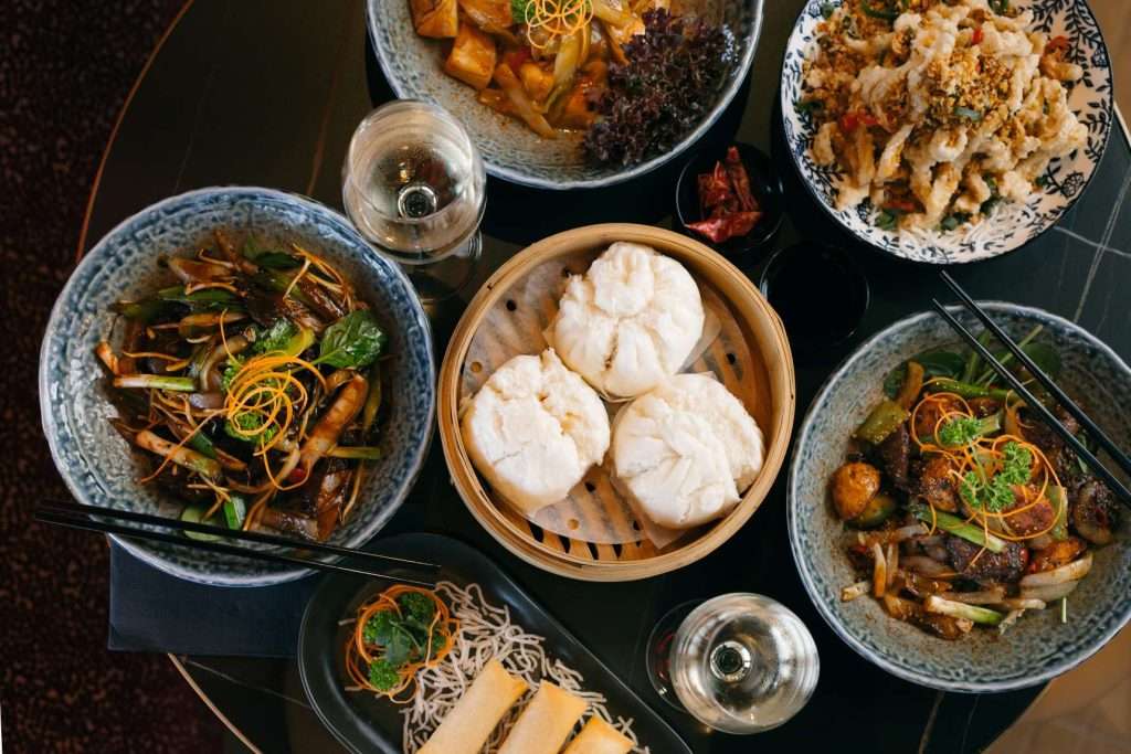 Overhead view of Asian-inspired dishes at Wok O’Clock Cairns, including steamed bao buns, stir-fried vegetables, crispy bites and spring rolls with glasses of white wine.